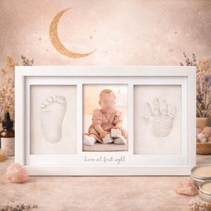 May include: White framed baby keepsake with a baby's foot and hand impressions, and a photo of a baby in a pink dress. The text "Love at first sight" is printed below the photo. The background is a soft, neutral colour.