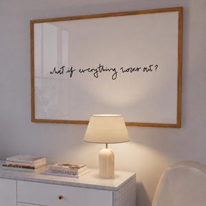 Op de afbeelding: Een rechthoekig ingelijst kunstwerk aan de muur met een lichthouten lijst en een witte achtergrond, voorzien van de zwarte cursieve tekst "what if everything works out?" boven een tafellamp en een witte kast met gestapelde boeken.