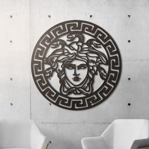 Puede incluir: Una pieza de arte mural de metal marrón oscuro que presenta la cabeza de Medusa dentro de un marco circular con un diseño de clave griega. La obra de arte está montada en una pared de hormigón, con dos sillones blancos y una mesa auxiliar dorada en primer plano.