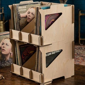 May include: A two-tier light-colored wooden vinyl record storage rack with triangular side cutouts. The rack holds multiple vinyl records, including David Bowie's Hunky Dory, The Grateful Dead, and John Coltrane's A Love Supreme, organized in slots.