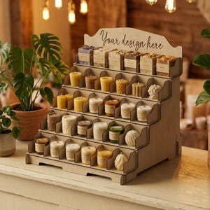 May include: A tiered wooden display stand with five shelves holding various small candles and soaps. The top header features the text "Your design here" in a cursive font. The stand is constructed from light-colored plywood with visible slots.