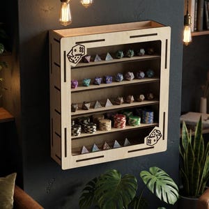 May include: A light-coloured wooden wall-mounted display shelf featuring five horizontal tiers. The frame includes laser-cut dice icons in the top-left and bottom-right corners. The shelves hold various multi-sided gaming dice and stacks of circular gaming chips.