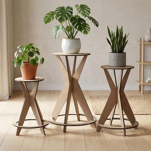 May include: Three light-coloured wooden plant stands with X-shaped legs and circular bases. The stands vary in height and support three potted plants: a small green plant in a terracotta pot, a monstera in a white pot, and a snake plant in a grey pot.