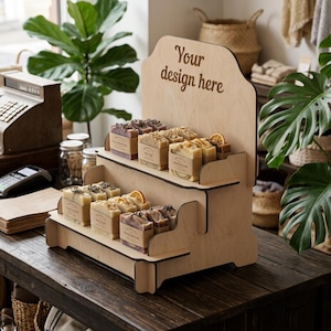 Könnte beinhalten: Ein zweistufiger Präsentationsständer aus hellem Holz mit einer hohen Rückwand, in die der Text Your design here eingraviert ist. Der Ständer enthält mehrere rechteckige Seifenstücke, die mit Papierbanderolen umwickelt und auf den Regalen angeordnet sind.