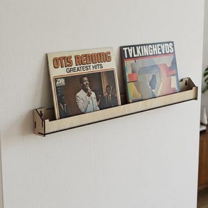 Könnte beinhalten: Ein helles Wandregal aus Holz, das zwei Schallplatten hält. Das linke Plattencover zeigt Otis Redding Greatest Hits, während das rechte Cover den Bandnamen Talking Heads in fetten, schwarzen Buchstaben auf der Vorderseite aufweist.