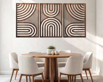 May include: Three-piece wooden wall art with a geometric design of arches and lines. The art is a dark brown color and is mounted on a white wall above a round wooden dining table and chairs. The room has a modern, minimalist aesthetic.
