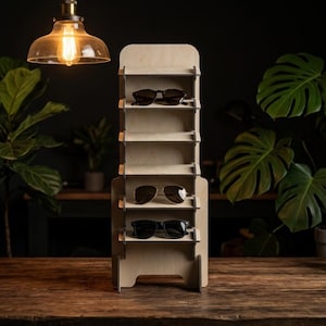 May include: A vertical light-colored wooden display stand with five horizontal shelves designed for eyewear. The stand holds three pairs of sunglasses, including dark-rimmed frames and aviator-style glasses, positioned on the top, middle, and bottom shelves.