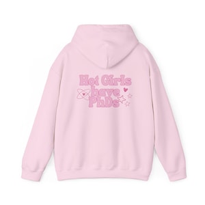 Pode incluir: Um moletom rosa claro com o texto "Hot Girls have PhDs" em letras rosa bolha. O design inclui corações, estrelas e um símbolo de átomo. O moletom tem capuz e um ajuste relaxado.