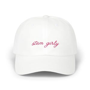 May include: A white baseball cap featuring the text "stem girly" embroidered in a dark pink cursive script across the front panel. The hat has a structured crown, a curved brim, and a button at the top center, providing a clean and simple design.