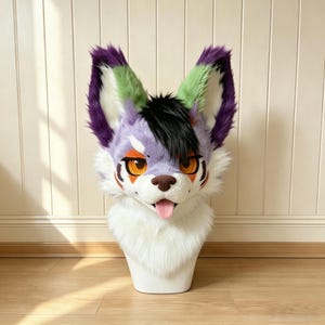 Op de afbeelding: Een imitatiebont dierenkopmasker met een lavendelkleurig en wit gezicht en grote puntige oren met paarse en groene uiteinden. Het masker heeft feloranje ogen, zwarte markeringen rond de ogen, een bruine neus en een roze tong die uit de mond steekt.