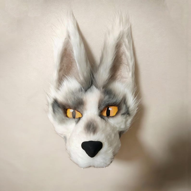May include: A faux fur animal mask featuring white and grey synthetic fur, tall pointed ears with light grey inner detailing, and bright orange eyes with black pupils. The mask has a solid black nose and a textured, realistic animal face shape.
