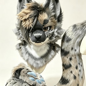 May include: A faux fur animal head mask with gray, white, and tan spotted patterns, featuring long pointed ears, a tuft of tan hair, and brown eyes. Included are matching gray and tan spotted paw gloves with blue pads and a long spotted tail.