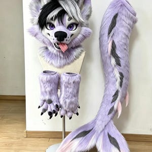 Könnte beinhalten: Ein Kunstfell-Tierkostümset bestehend aus einer lavendelfarbenen und weißen Wolfsmaske mit schwarzem Haar, violetten Augen und einer rosa Zunge. Das Set enthält zwei passende lavendelfarbene Pfotenhandschuhe mit schwarzen Krallen und einen langen Schwanz.