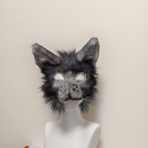 May include: A gray and black faux fur animal mask featuring pointed ears, a textured snout with dark spots, and two white fangs protruding from the lower jaw area. The mask has cutouts for the eyes and is displayed on a white mannequin head.