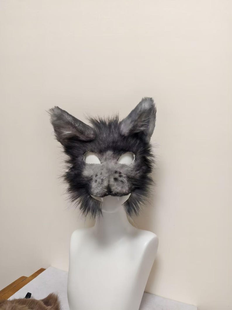 May include: A gray and black faux fur animal mask featuring pointed ears, a textured snout with dark spots, and two white fangs protruding from the lower jaw area. The mask has cutouts for the eyes and is displayed on a white mannequin head.