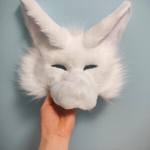May include: A white faux fur animal mask featuring tall pointed ears, a prominent snout, and narrow eye openings. The mask is covered in soft, long-pile white synthetic fur, creating a textured appearance with a distinct animal-like facial structure.