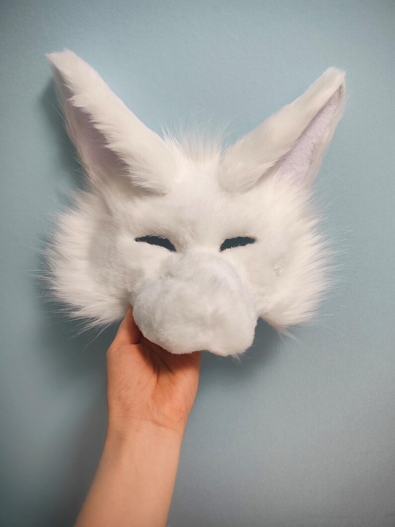 May include: A white faux fur animal mask featuring tall pointed ears, a prominent snout, and narrow eye openings. The mask is covered in soft, long-pile white synthetic fur, creating a textured appearance with a distinct animal-like facial structure.