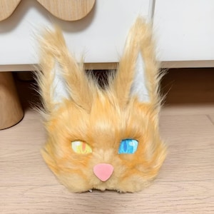 May include: A faux fur animal mask featuring long, pointed ears with white inner detailing, a soft orange coat, a small pink nose, and two distinct eyes with a yellow iris on the left and a blue iris on the right, both showing a grid-like texture.