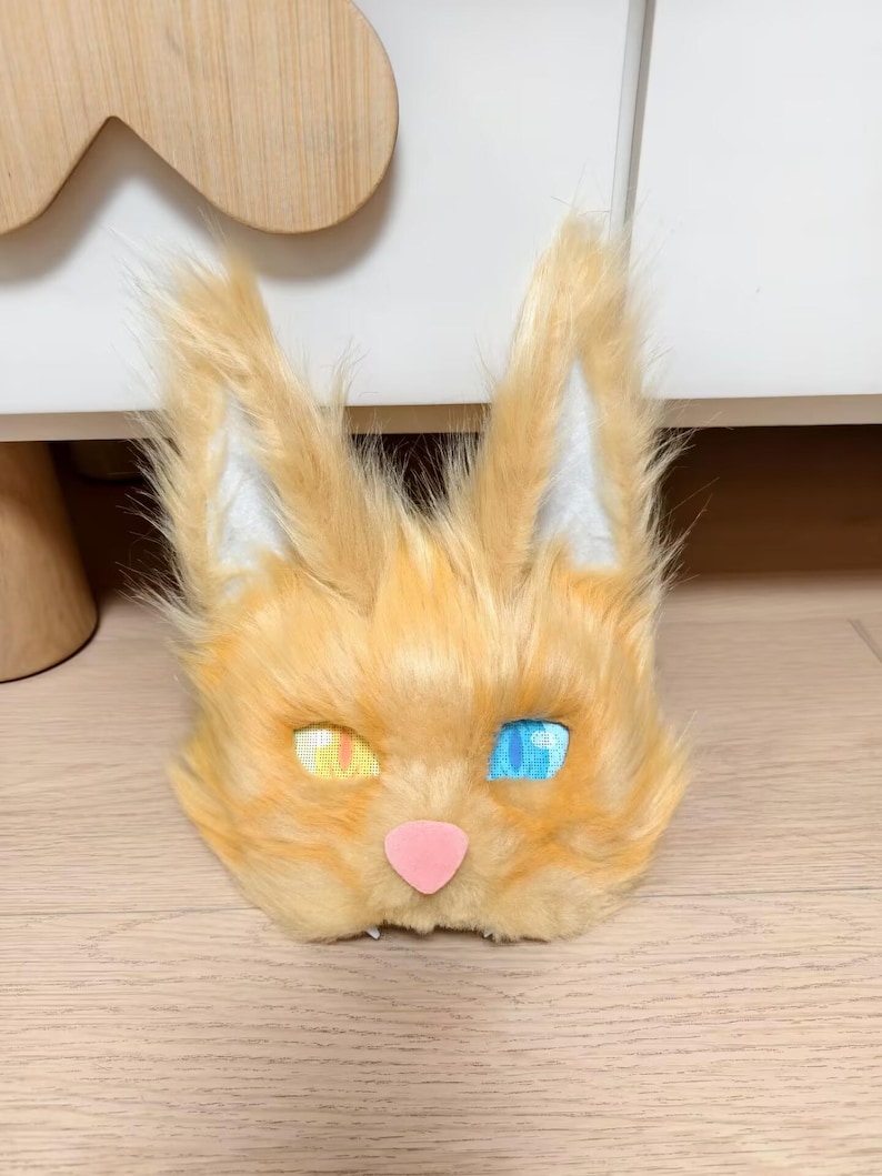 May include: A faux fur animal mask featuring long, pointed ears with white inner detailing, a soft orange coat, a small pink nose, and two distinct eyes with a yellow iris on the left and a blue iris on the right, both showing a grid-like texture.