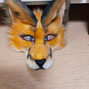 May include: A faux fur animal mask featuring orange and black fur, pointed ears with dark tips, and a black nose. The eyes are covered with a fine mesh material, and the overall design mimics the facial features of a fox or similar wild feline.