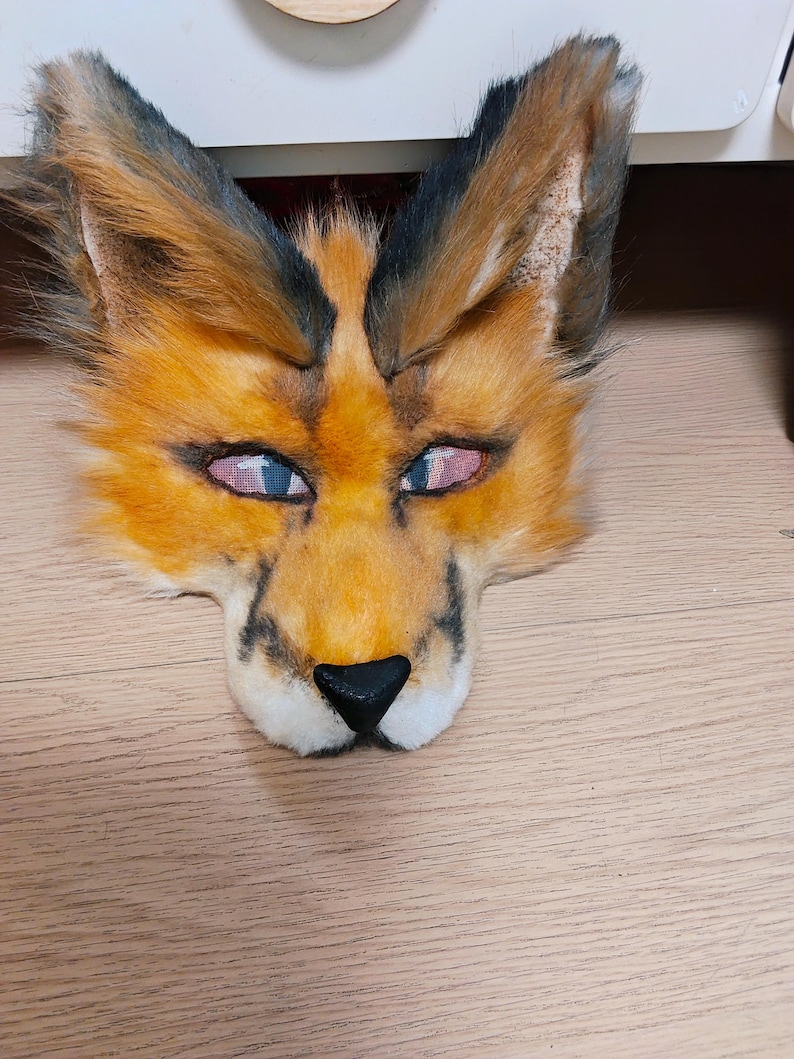 May include: A faux fur animal mask featuring orange and black fur, pointed ears with dark tips, and a black nose. The eyes are covered with a fine mesh material, and the overall design mimics the facial features of a fox or similar wild feline.