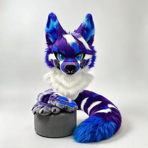 Puede incluir: Una máscara de cabeza de animal de peluche con piel sintética en colores morado, blanco y azul brillante. La máscara tiene grandes ojos azules expresivos, orejas puntiagudas con detalles internos en azul y una larga cola a rayas a juego.