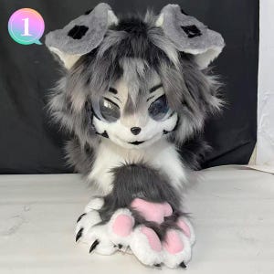 Puede incluir: Una cabeza de fursuit peluda, gris y blanca, con ojos grandes y expresivos. Las orejas son grises con detalles negros, y las patas son blancas con almohadillas rosas y garras negras. El número "1" está en un círculo rosa y verde en la esquina superior izquierda.