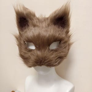 May include: A brown faux fur animal mask featuring pointed ears and almond-shaped eye openings. The mask is covered in soft, textured brown synthetic fibers that mimic animal fur, with a rounded snout area and a wide, expressive feline-like shape.