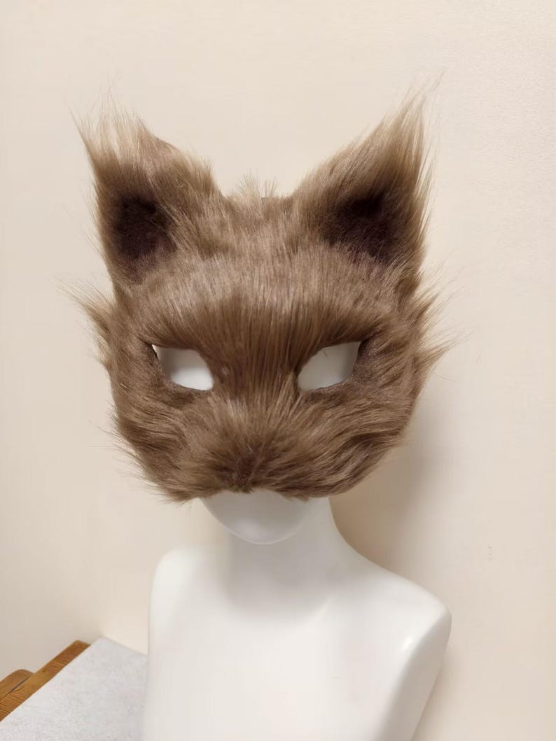 May include: A brown faux fur animal mask featuring pointed ears and almond-shaped eye openings. The mask is covered in soft, textured brown synthetic fibers that mimic animal fur, with a rounded snout area and a wide, expressive feline-like shape.