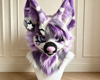 Könnte beinhalten: Eine Plüsch-Tierkopfmaske mit langen, spitzen Ohren aus violettem, weißem und grauem Kunstfell. Die Maske hat große, ausdrucksstarke Augen mit violetten Iris, eine rosa Nase und einen kleinen silbernen Nasenring. Zwei sternförmige Anhänger hängen an einem Ohr.