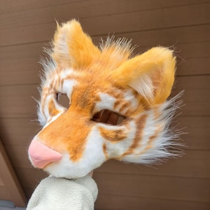 May include: A tiger-themed animal mask crafted from faux fur in shades of orange and white, featuring pointed ears, a smooth pink nose, and cutouts for the eyes. The mask displays a striped pattern across the forehead and cheeks with long fur.