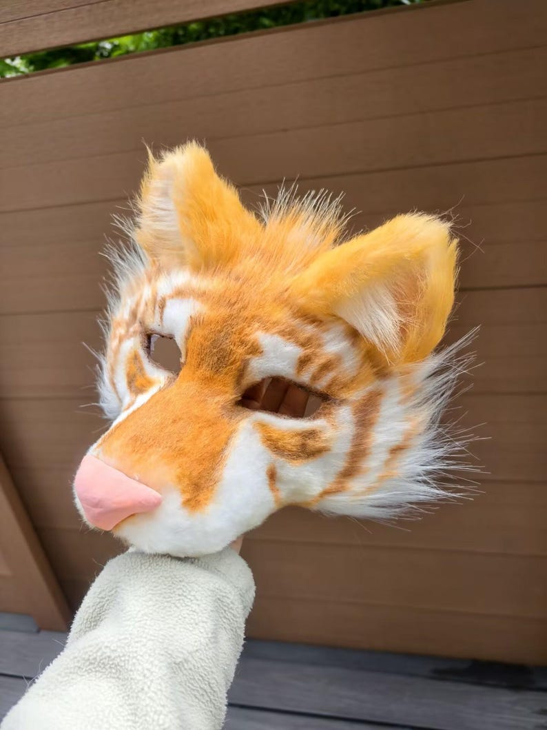May include: A tiger-themed animal mask crafted from faux fur in shades of orange and white, featuring pointed ears, a smooth pink nose, and cutouts for the eyes. The mask displays a striped pattern across the forehead and cheeks with long fur.