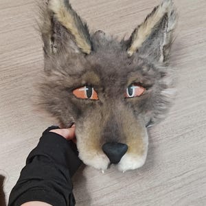 May include: A gray and tan faux fur wolf mask featuring pointed ears, orange eyes with black pupils, and a black nose. The mask has a textured, shaggy appearance with lighter fur accents around the ears and muzzle, designed to cover the face.