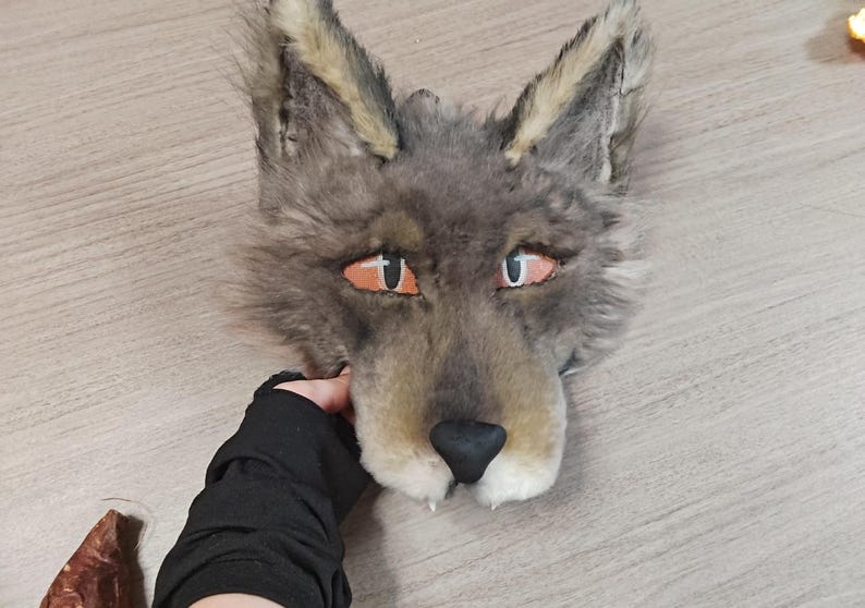 May include: A gray and tan faux fur wolf mask featuring pointed ears, orange eyes with black pupils, and a black nose. The mask has a textured, shaggy appearance with lighter fur accents around the ears and muzzle, designed to cover the face.