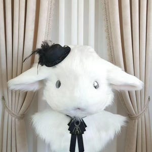 May include: A white rabbit head with long ears, a small black hat with a feather, and a black bow tie. The rabbit head is made of fluffy material and is displayed on a white mannequin head. The background features beige curtains.