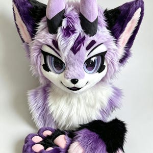 May include: A plush animal head mask featuring long lavender and white faux fur, large expressive purple eyes, black markings, and two upright horns. The mask is accompanied by a pair of matching purple, black, and pink paw-shaped hand covers.