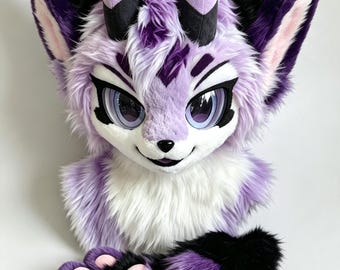Könnte beinhalten: Eine Plüsch-Tierkopfmaske mit langem lavendelfarbenem und weißem Kunstfell, großen ausdrucksstarken violetten Augen, schwarzen Markierungen und zwei aufrechten Hörnern. Die Maske wird von einem Paar passender pfotenförmiger Handschuhe ergänzt.