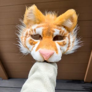 May include: A tiger mask crafted from faux fur in shades of orange and white, featuring pointed ears, dark brown striped markings, and a smooth, light pink nose. The mask has two eye openings and a textured, fluffy appearance throughout the design.