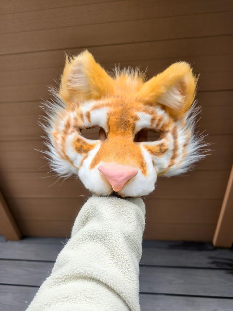 May include: A tiger mask crafted from faux fur in shades of orange and white, featuring pointed ears, dark brown striped markings, and a smooth, light pink nose. The mask has two eye openings and a textured, fluffy appearance throughout the design.