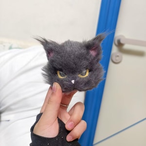 May include: A handcrafted brooch shaped like a fluffy dark gray cat head with pointed ears, textured fur, yellow eyes with a grid pattern, and a small white nose. The cat head features a stern expression and is made of soft, fuzzy material.