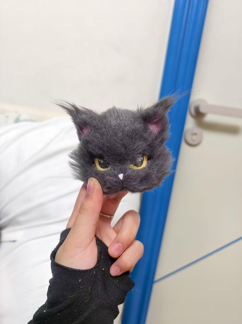 May include: A handcrafted brooch shaped like a fluffy dark gray cat head with pointed ears, textured fur, yellow eyes with a grid pattern, and a small white nose. The cat head features a stern expression and is made of soft, fuzzy material.