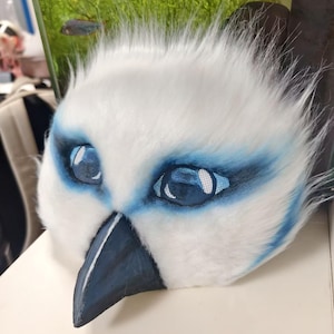 May include: A white faux fur mask featuring a pointed dark gray beak and large, expressive blue eyes with mesh detailing. The mask is accented with blue airbrushed markings around the eye sockets and has a soft, textured appearance throughout.