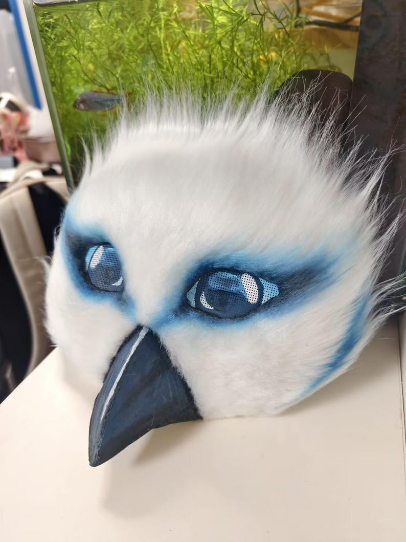 May include: A white faux fur mask featuring a pointed dark gray beak and large, expressive blue eyes with mesh detailing. The mask is accented with blue airbrushed markings around the eye sockets and has a soft, textured appearance throughout.
