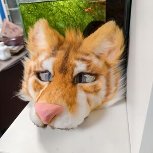 May include: A feline-inspired mask featuring orange and white faux fur with dark brown stripes. The mask has pointed ears, a smooth pink nose, and mesh-covered eye openings with visible pupils, designed to cover the head and face area.