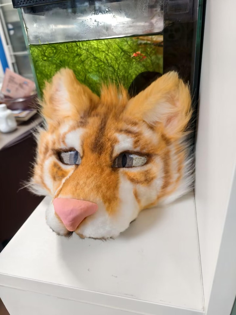 May include: A feline-inspired mask featuring orange and white faux fur with dark brown stripes. The mask has pointed ears, a smooth pink nose, and mesh-covered eye openings with visible pupils, designed to cover the head and face area.