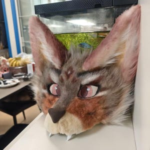 May include: A faux fur animal head mask featuring tall, pointed ears with pinkish-tan inner fur, a grey and brown patterned face, large mesh eyes with pink irises, a dark brown nose, and two white fangs protruding from the cream-colored muzzle.