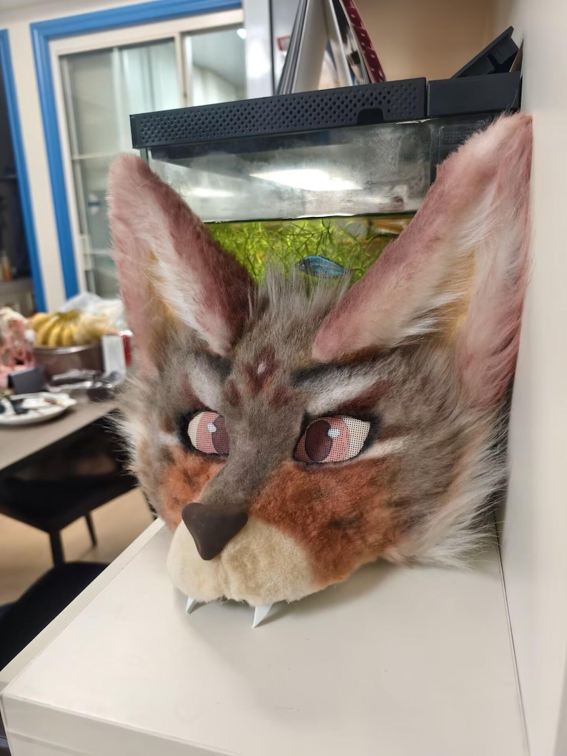 May include: A faux fur animal head mask featuring tall, pointed ears with pinkish-tan inner fur, a grey and brown patterned face, large mesh eyes with pink irises, a dark brown nose, and two white fangs protruding from the cream-colored muzzle.