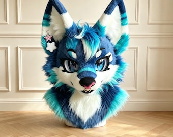 Könnte beinhalten: Eine Plüsch-Tierkopfmaske mit langen, spitzen Ohren aus blauem, weißem und türkisfarbenem Fell. Das Gesicht hat große blaue Augen, eine schwarze Nase, eine kleine rosa Zunge und einen goldenen Nasenring. Ein rosa und ein weißer Stern hängen am linken Ohr.