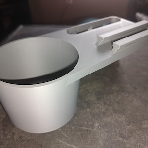 May include: A light gray 3D printed cup holder attachment featuring a cylindrical vessel for holding drinks and an integrated flat tray section with two parallel raised ridges designed for securing small items or mobile devices.