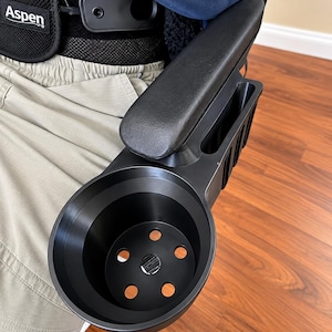 May include: A black plastic wheelchair armrest attachment featuring a circular cup holder with six drainage holes at the base and an integrated rectangular storage compartment for small items, mounted to the side of a padded armrest.